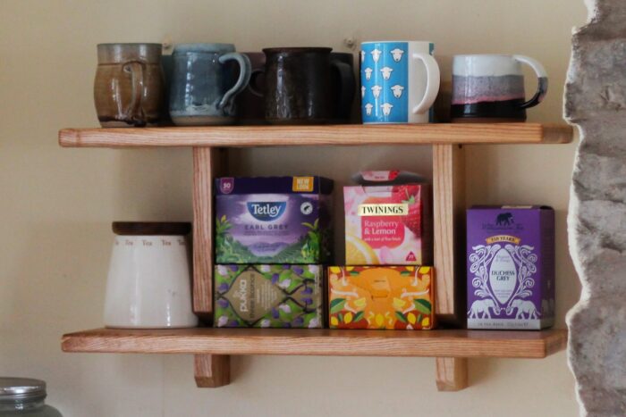 Two wooden shelves containing boxes of tea, a jar of tea and a variety of mugs